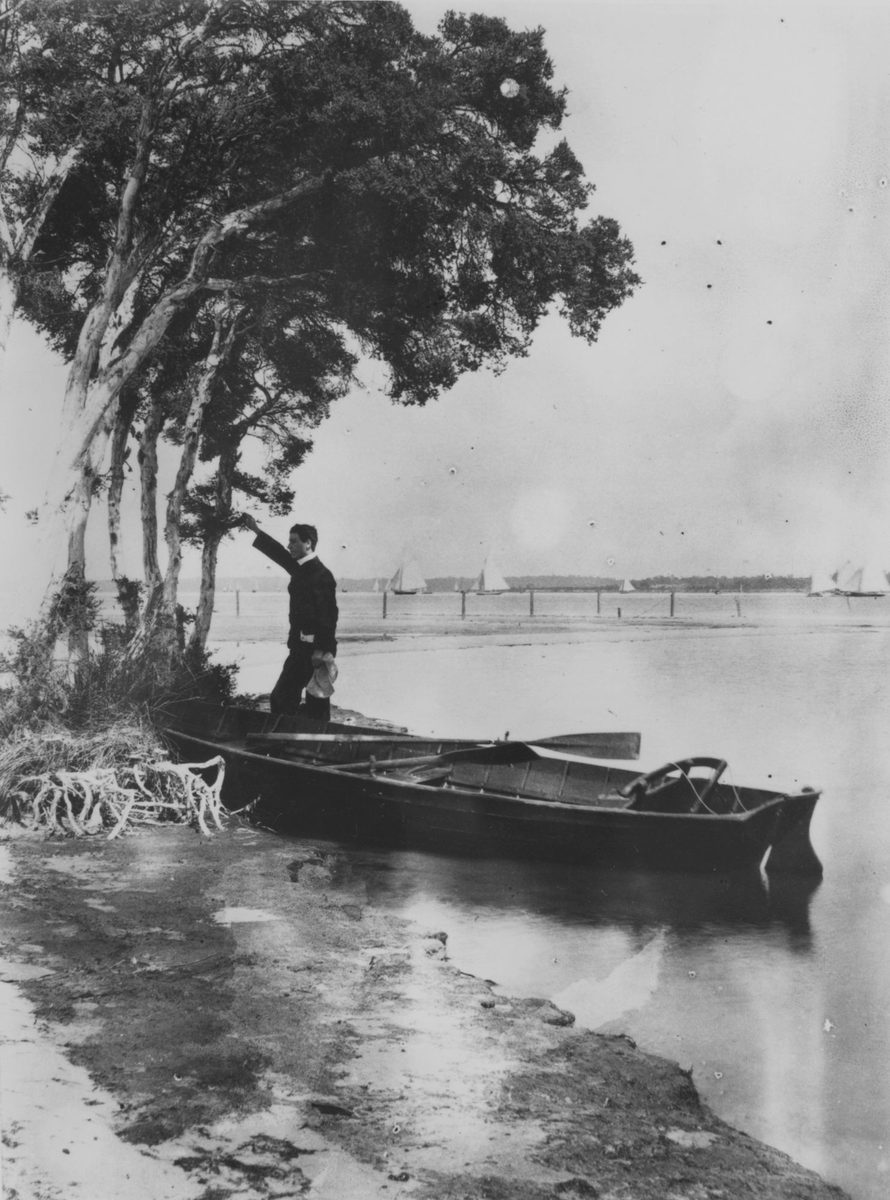 Man standing on a boat tied to a tree at the edge of Brisbane River, 1868