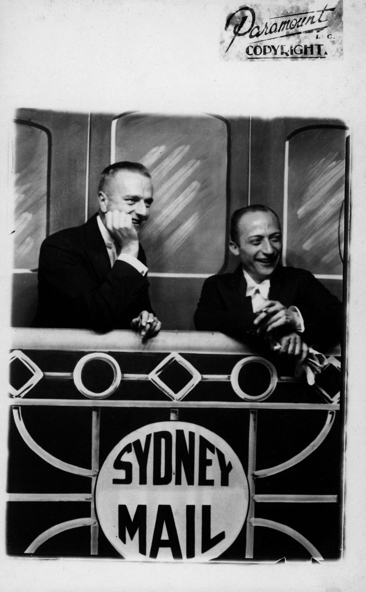 Two gentlemen in dinner suits posing with a backdrop of the 'Sydney Mail train' at the Trocadero