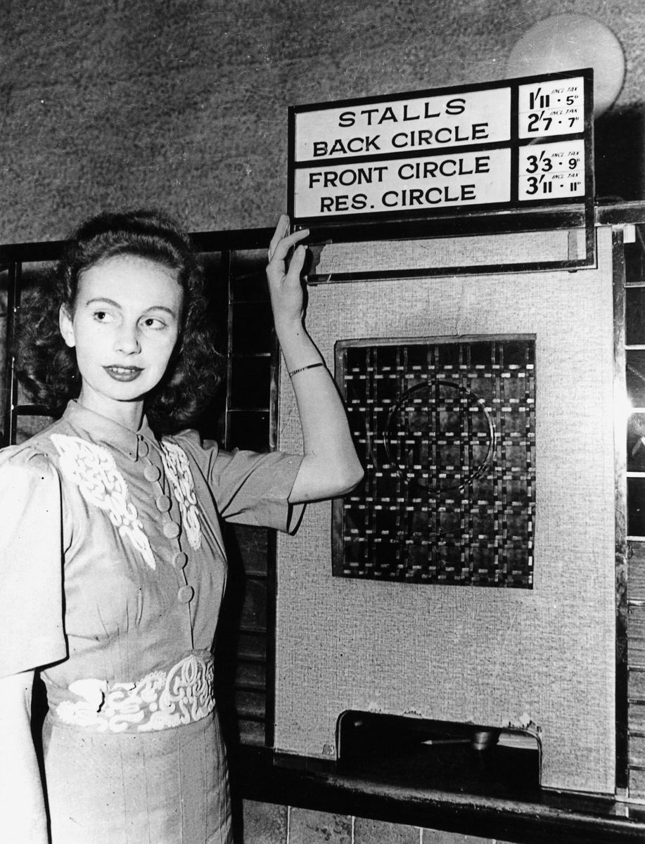 Young lady displaying noticeboard with new prices for entertainment, including tax, Brisbane, 1942